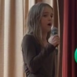 Close up of a 10-year-old girl a she sings into a mic. Nearby to one side is a cluster of green and yellow balloons.