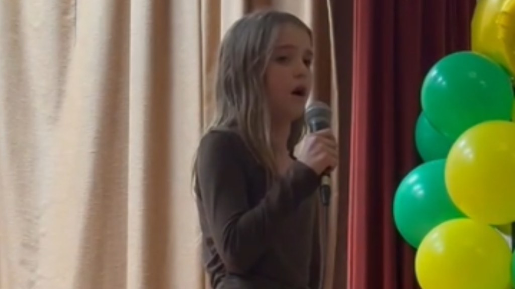 Close up of a 10-year-old girl a she sings into a mic. Nearby to one side is a cluster of green and yellow balloons.